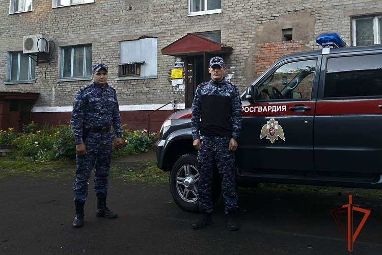 Двое сотрудников Росгвардии стоят рядом со служебным автомобилем. На заднем плане — многоэтажный кирпичный жилой дом