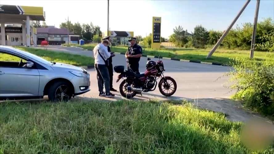 двое сотрудников Госавтоинспекции беседуют с мотоциклистом, рядом стоит красный мотоцикл, фоном - зелень и сельская местность