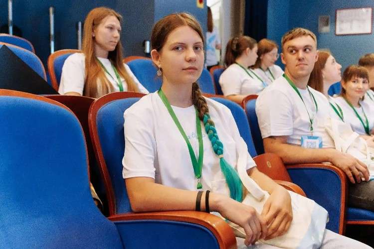 Фотография молодой девушки-подростка, она сидит в одном из кресел в актовом зале, её волосы заплетены в косу, кончик косы - синий.