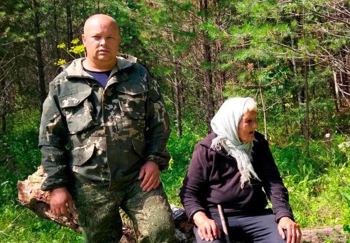 Пожилая женщина сидит на пеньке. Рядом стоит мужчина