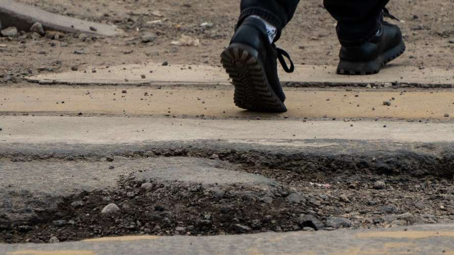 Ноги человека в черных ботинках идут по разбитому асфальту. На переднем плане — крупная выбоина с осыпавшимися краями и щебнем.