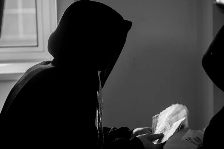 Черно-белое фото: силуэт человека в капюшоне, сидящего у окна. В руках он держит веер из денежных купюр и перебирает их. Рядом справа виден край силуэта другого человека.