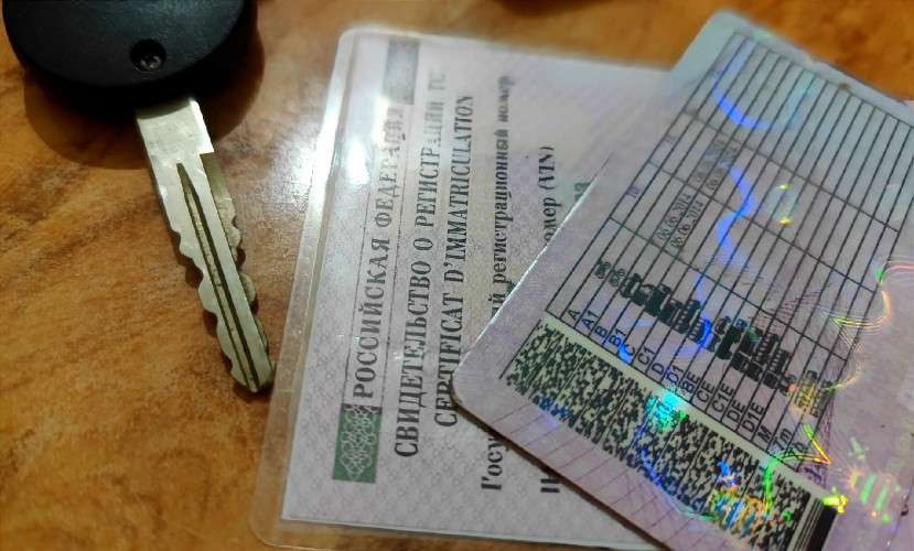 На деревянной поверхности лежат автомобильный ключ с черным брелоком и два документа: свидетельство о регистрации транспортного средства (СТС) и водительское удостоверение с голографическими элементами и таблицей категорий.