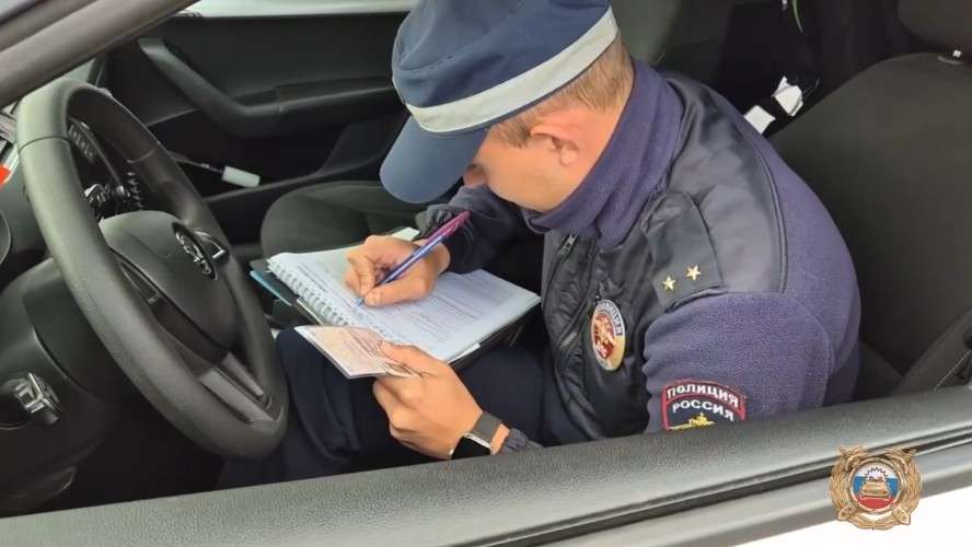 Сотрудник полиции в темно-синей форме сидит за рулем автомобиля и заполняет документы ручкой, опираясь на блокнот и бумажник. На левом плече видны две звездочки.