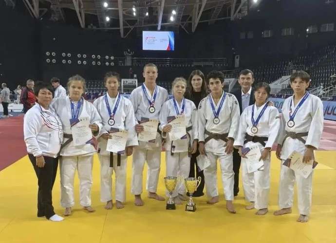 Группа юных спортсменов в белых кимоно (с поясами разного цвета) и их тренеры стоят на желтом татами в спортивном зале. У ребят на шеях висят медали, в руках — грамоты. Перед ними на полу стоят два золотистых кубка.
