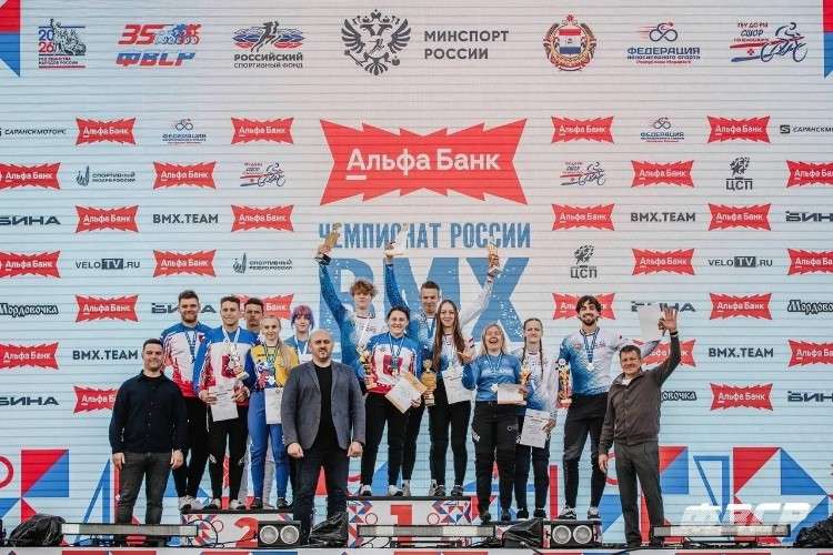 Группа спортсменов и официальных лиц на пьедестале во время награждения. Спортсмены одеты в яркую форму, у многих на шее медали, в руках кубки и грамоты. За ними баннер с надписями «ЧЕМПИОНАТ РОССИИ BMX» и логотипами спонсоров.