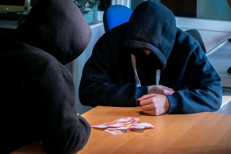 Два мошенника в черных капюшонах сидят за деревянным столом. Их лица скрыты. Перед ними лежит небольшая пачка российских денежных купюр. Атмосфера скрытная, напоминающая сделку.