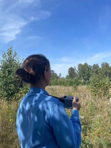 Женщина в голубой рубашке стоит спиной к камере на заросшем травой поле. В руках она держит устройство пульт управления дроном, направив его в сторону леса.