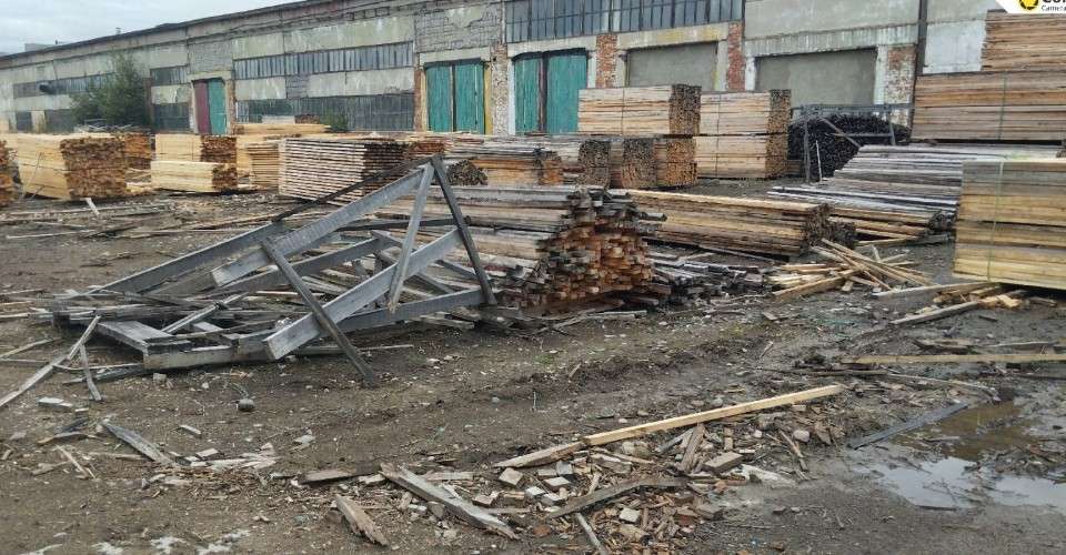 Территория лесопилки или склада пиломатериалов. На переднем плане на грязной земле лежат обломки досок, деревянные щепки и груда металлических ферм. Позади расположены аккуратные штабеля свежих досок.