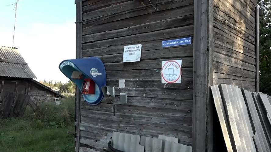 На стене старого деревянного дома установлен синий уличный таксофон с красной трубкой.