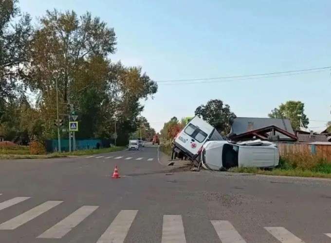 Дорожно-транспортное происшествие на перекрестке. Белый автомобиль скорой помощи лежит на боку на обочине. Рядом стоит оранжевый дорожный конус. На заднем плане видны пешеходный переход, светофор, деревья и дома.