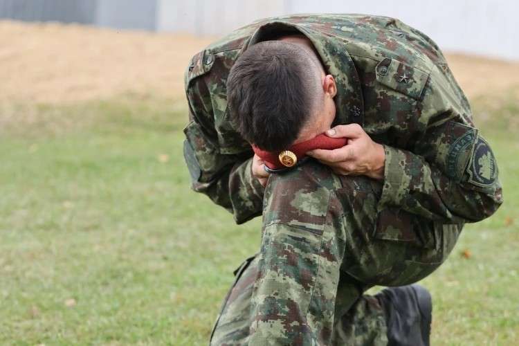 Военнослужащий в камуфляжной форме с шевроном на рукаве стоит на колене, низко опустив голову. Обеими руками он прижимает к лицу красную фуражку с золотой кокардой. Лицо солдата скрыто.