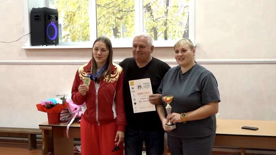 Трое людей позируют для фото в помещении. Слева девушка в красном спортивном костюме с золотой медалью на шее. В центре мужчина в черной футболке держит диплом. Справа женщина в серой футболке с золотым кубком в руках.