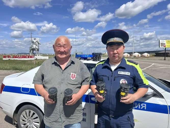 Сотрудник ГИБДД и водитель стоят на фоне машины полиции. У них в руках стаканчики с кофе.