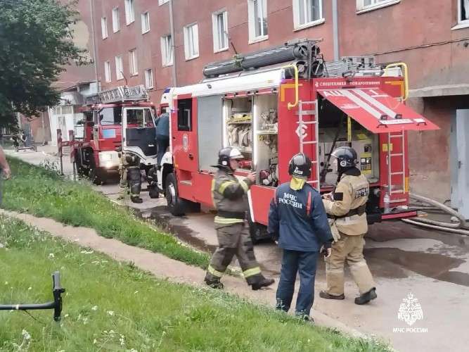 Вид с улицы на кирпичное здание, видно первые три этажа, на тротуаре стоят две пожарные машины и стоит группа пожарных, воздух немного непрозрачный из-за дыма, но пламя не видно