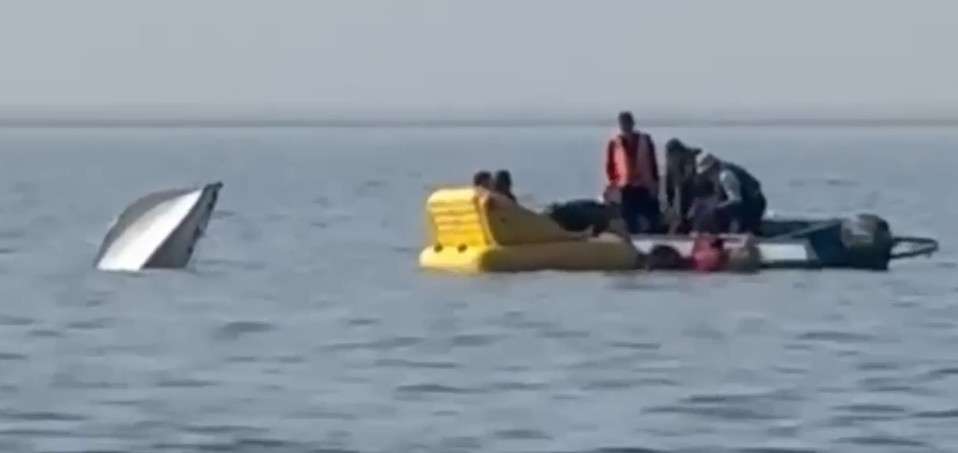 На воде затонула белая моторная лодка — виден только ее нос, торчащий из воды. Рядом люди на желтой резиновой лодке и небольшой моторной лодке занимаются спасением.