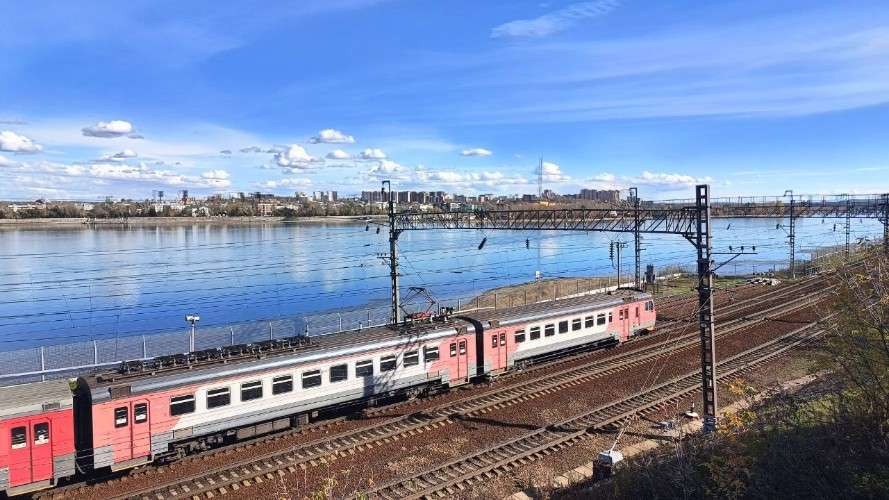 Поезд движется по путям прямо вдоль берега широкой голубой реки. На противоположном берегу видна панорама города с высотными зданиями. День ясный, небо ярко-голубое с белыми облаками.