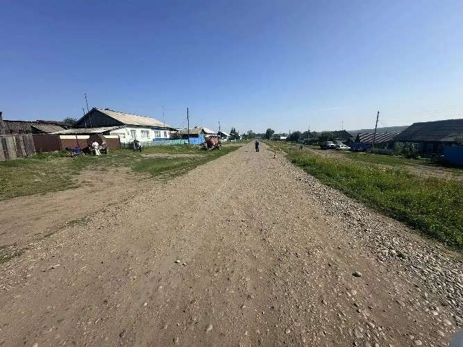 Широкая грунтовая дорога с камнями уходит вдаль. По центру дороги вдалеке идет человек, рядом стоит оранжевый конус. По бокам расположены одноэтажные сельские дома с заборами