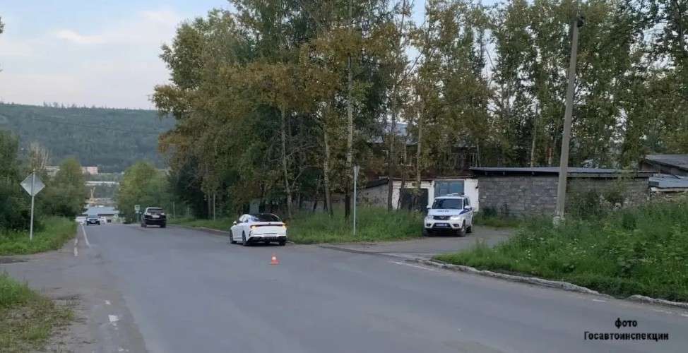 Дорога, ведущая вдаль. Слева стоит белая машина, рядом на дороге — оранжевый конус. Справа на обочине припаркован белый полицейский автомобиль. Вокруг — деревья, дома и лес на фоне.