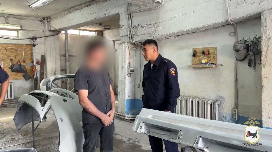 В помещении автосервиса или гаража беседуют двое мужчин. Слева стоит мужчина в темной одежде (лицо размыто), справа — сотрудник полиции в темно-синей форме с шевроном на рукаве.