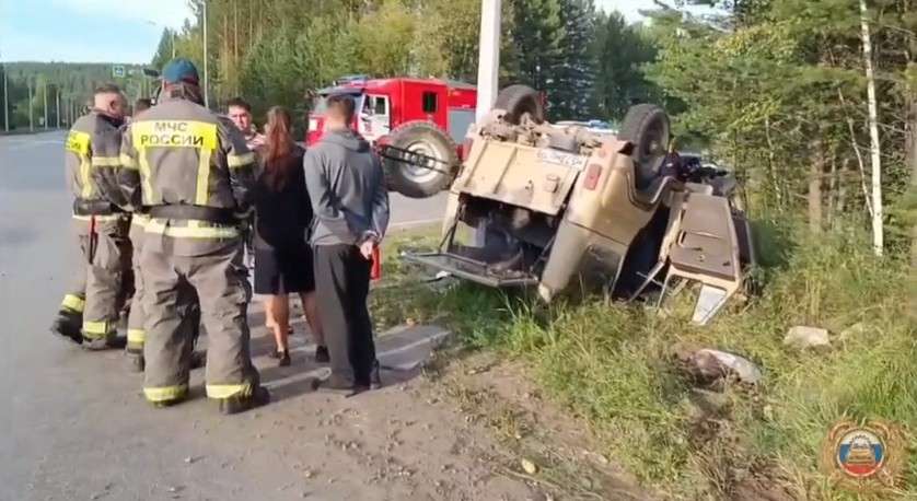 На обочине дороги перевернулся и лежит на боку автомобиль. Рядом стоят сотрудники МЧС России в защитной форме и несколько гражданских, которые наблюдают за происходящим. На заднем плане виден красный пожарный автомобиль.