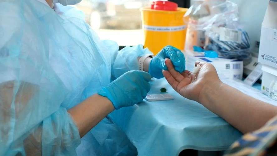 Медицинский работник в голубом защитном халате и перчатках берет кровь из пальца пациента. На столе лежит тест-кассета, рядом стоят контейнеры для анализов. Процедура происходит в светлом помещении.