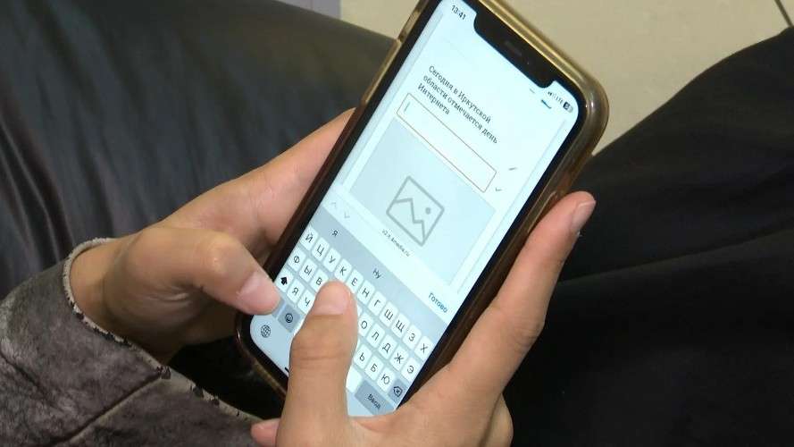 Руки человека держат смартфон в прозрачном чехле. На экране открыто приложение с текстом на русском: «Сегодня в Иркутской области отмечается день Интернета», ниже — пустое поле для ввода и место под картинку.