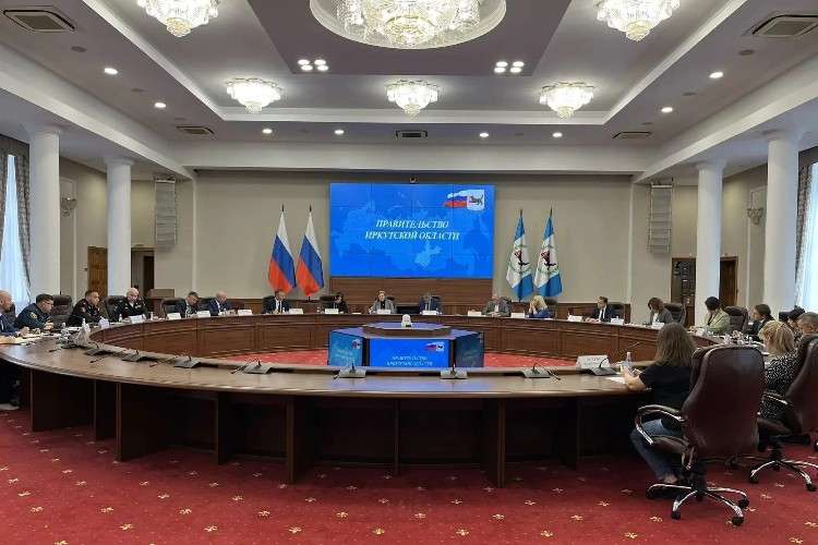 Официальное заседание в большом зале с колоннами. За длинным овальным столом сидят люди в деловых костюмах и форме. По бокам установлены флаги России и Иркутской области, потолок украшен хрустальными люстрами.
